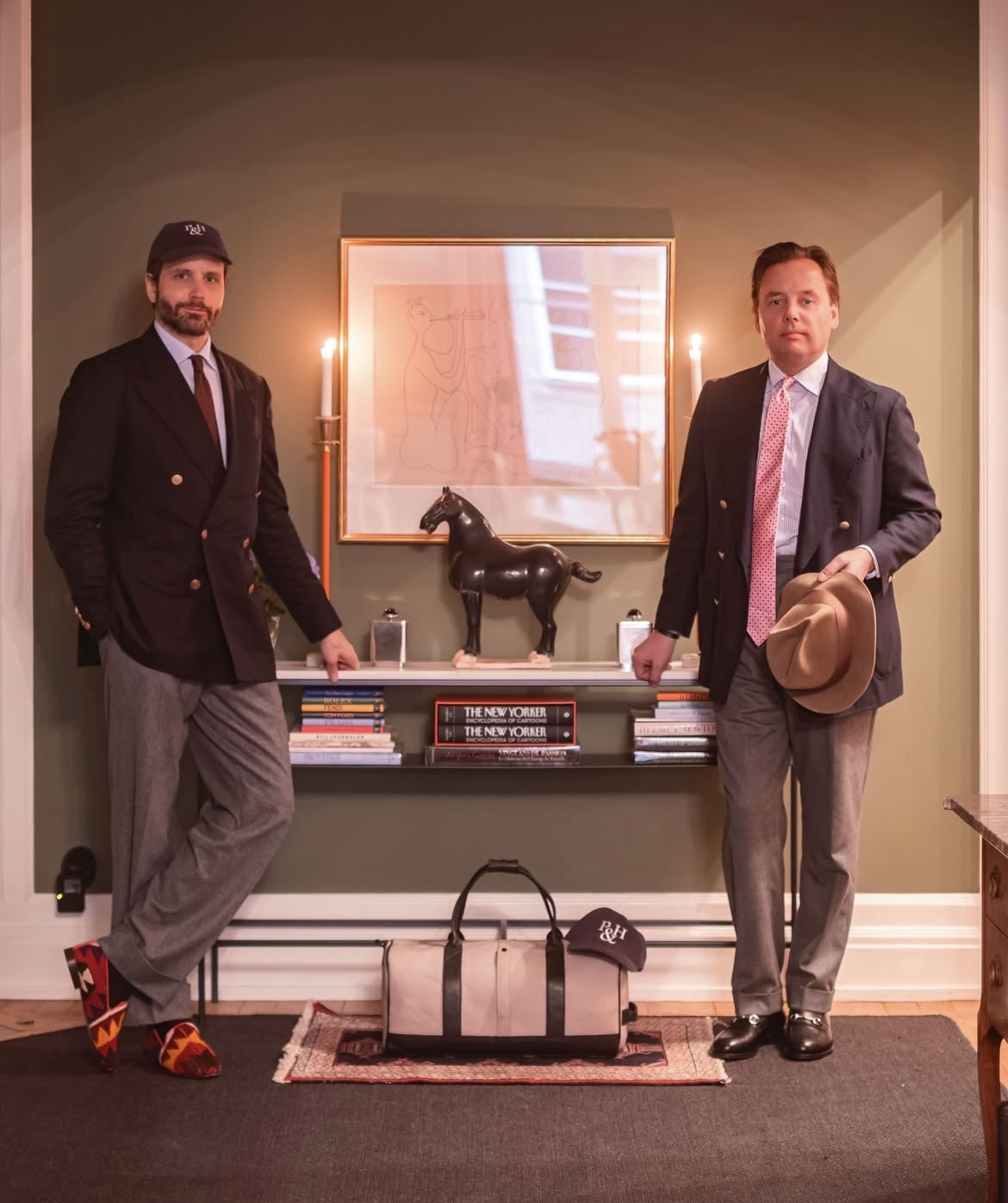Two men in blazers stand on either side of a wall-mounted shelf displaying books, a black horse sculpture, and a framed abstract artwork.