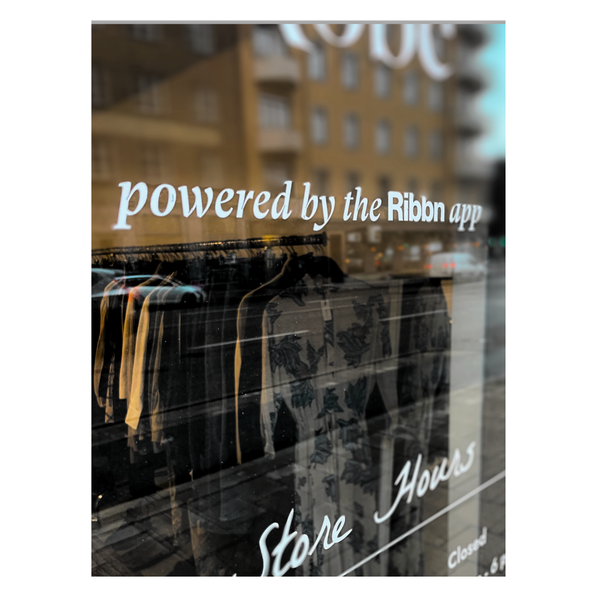 Window signage reading 'powered by the Ribbn app' with store hours visible, overlooking a street with buildings.