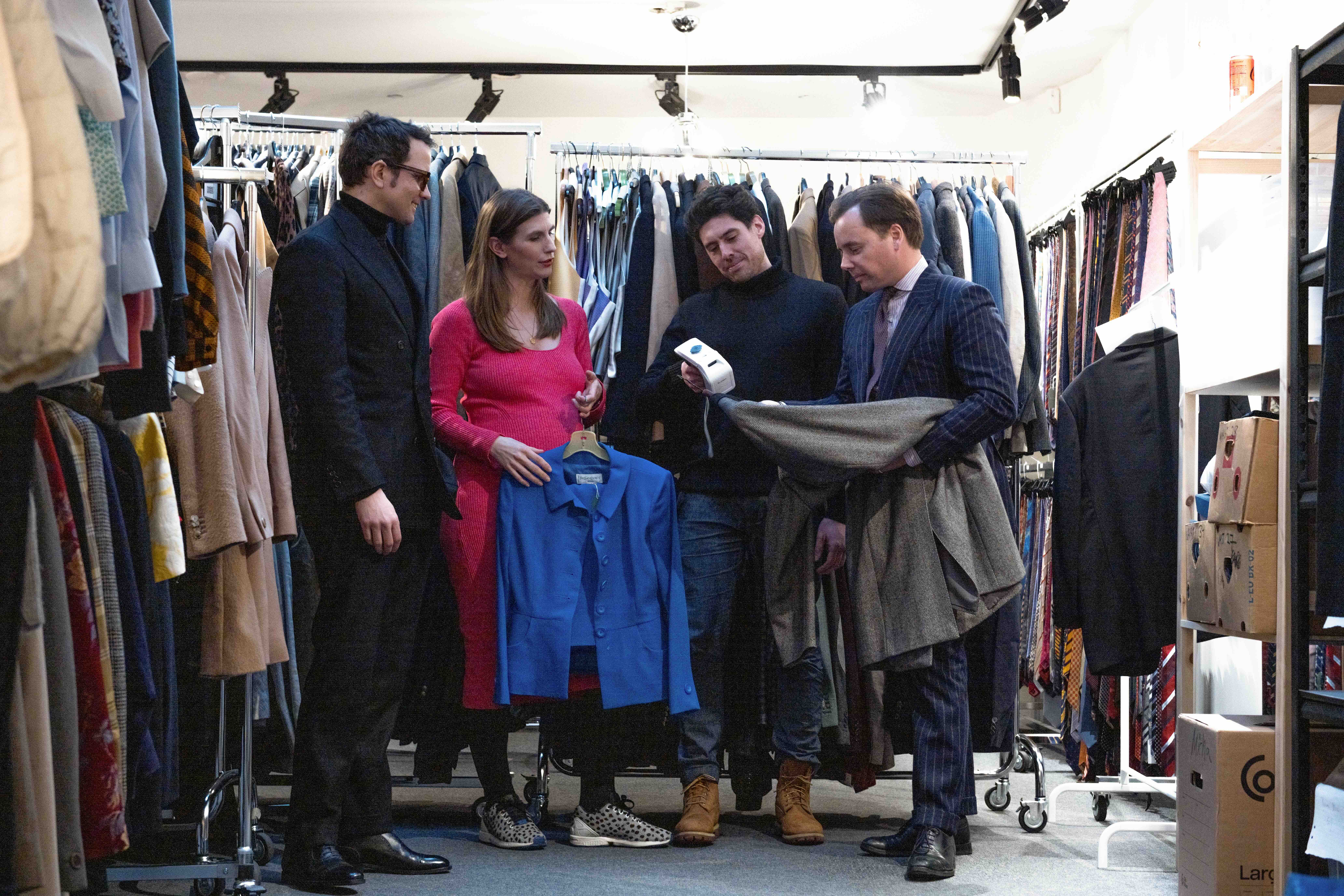 Four people standing among packed clothing rails in a stockroom, examining a blue jacket and a folded tweed garment while one scans a tag with a handheld device.